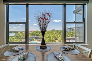 A dining table with placemats and a vase of red flowers, set against a large window with a view of a cityscape and greenery. - Harbor Towers 401 - Tropical Sands Accommodations (Siesta Key)