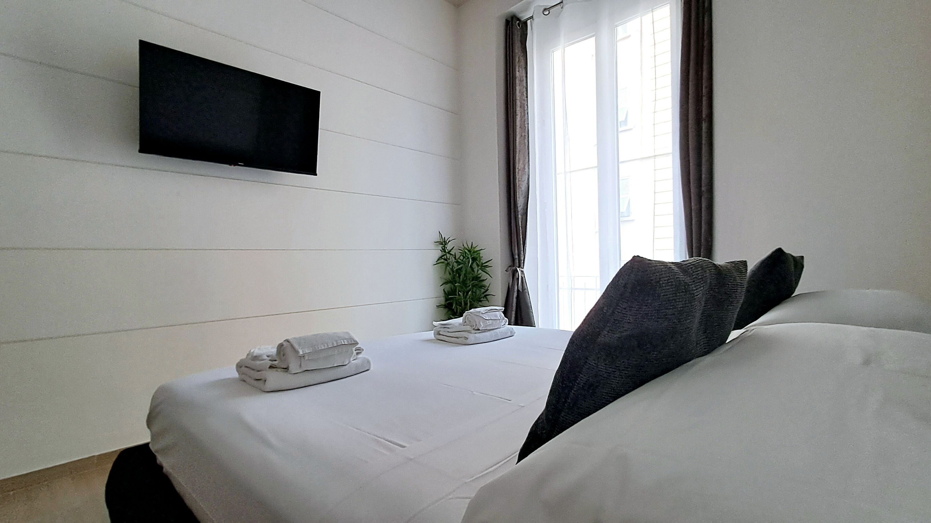 Habitación de hotel con cama, televisor montado en la pared, planta y ventana con cortinas.