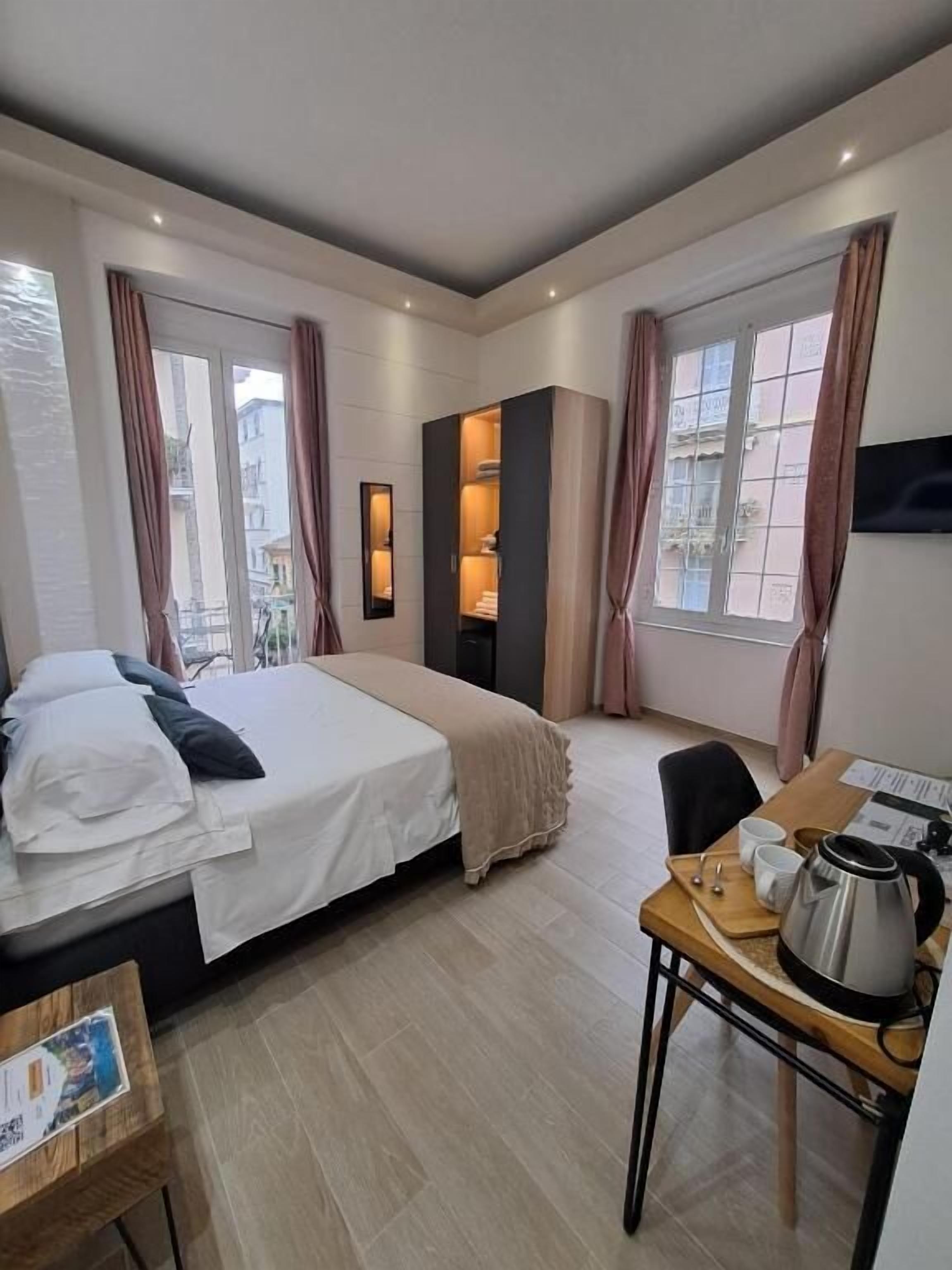 A hotel room with a bed, a desk with a kettle, and a view of buildings through the windows.