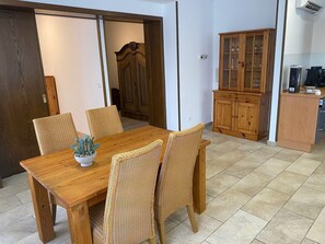 Ein Essbereich mit Holztisch und Korbstühlen, einem Schrank mit Glastüren sowie einer Küchenzeile mit Geräten.