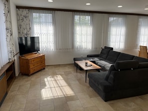 Ein Wohnzimmer mit einer Couch, einem Fernseher, einem Holzschrank und einem Fenster mit transparenten Vorhängen.