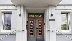 Entrada de un edificio con puertas dobles, un cartel que dice 'Ostecmann' arriba y una placa pequeña debajo de la puerta.