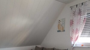 A loft-style bedroom with a sectional sofa, a large window with blinds, and a radiator. - Cozy vacation apartment in the countryside (Wertheim)