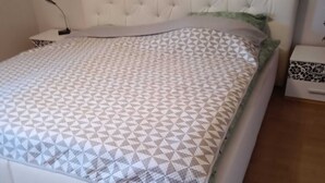 A bedroom with a white bed, a patterned bedspread, a tufted headboard, a bedside table with a patterned drawer, and a nightstand with a lamp. - Cozy vacation apartment in the countryside (Wertheim)