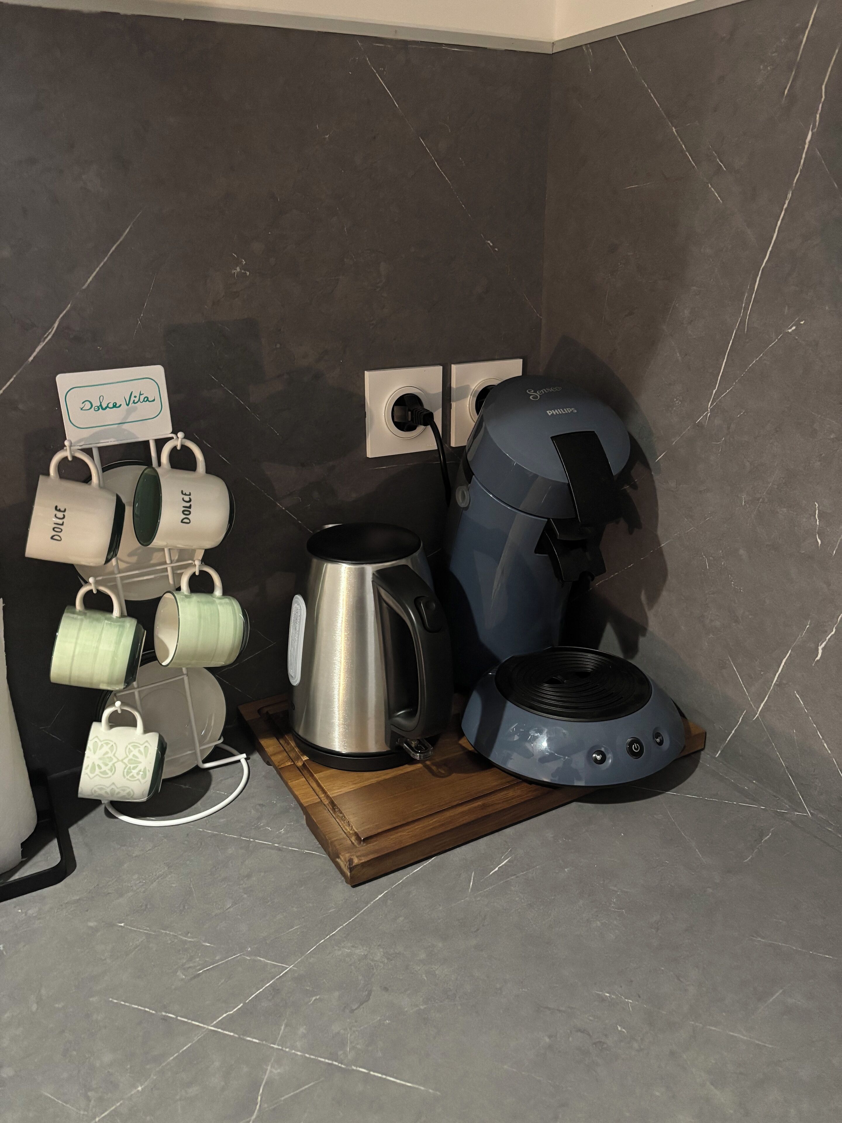 A kitchen area with a coffee maker, a kettle, and a mug rack with labeled mugs.