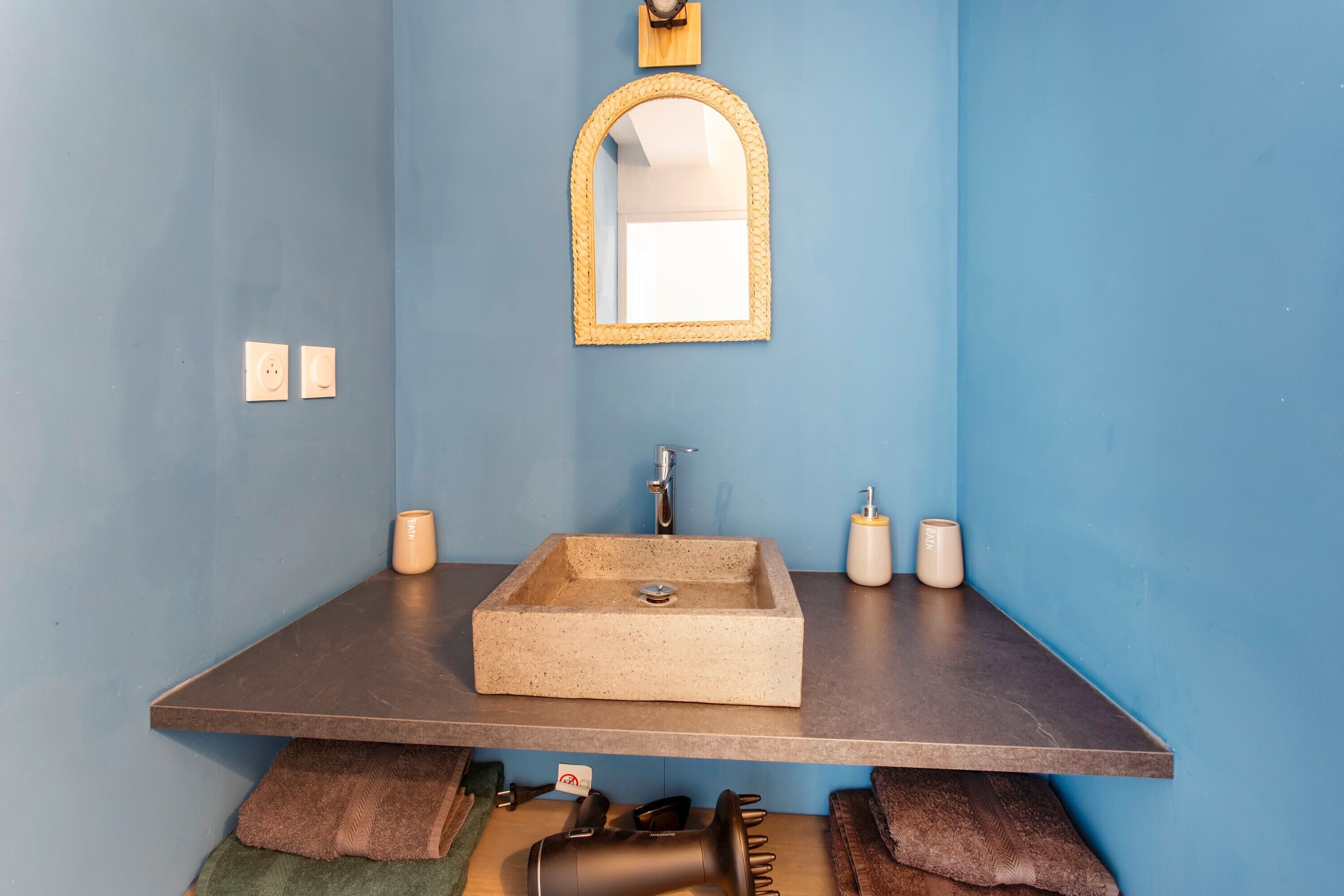 Une salle de bain avec un lavabo rectangulaire, un miroir doté d’un cadre tressé et deux bougies posées sur le comptoir.