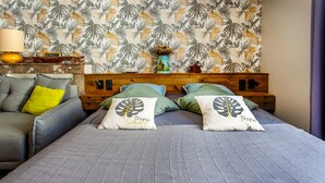 Ein Bett mit zwei Kissen mit der Aufschrift 'Tropic Summer', ein hölzernes Kopfteil, eine Lampe und eine Wand mit tropischem Blattmuster.