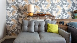Ein graues Sofa mit grau-gelben Kissen, ein Holztisch mit Lampe und einem kleinen Schild, und eine Wand mit tropischem Blattmuster.