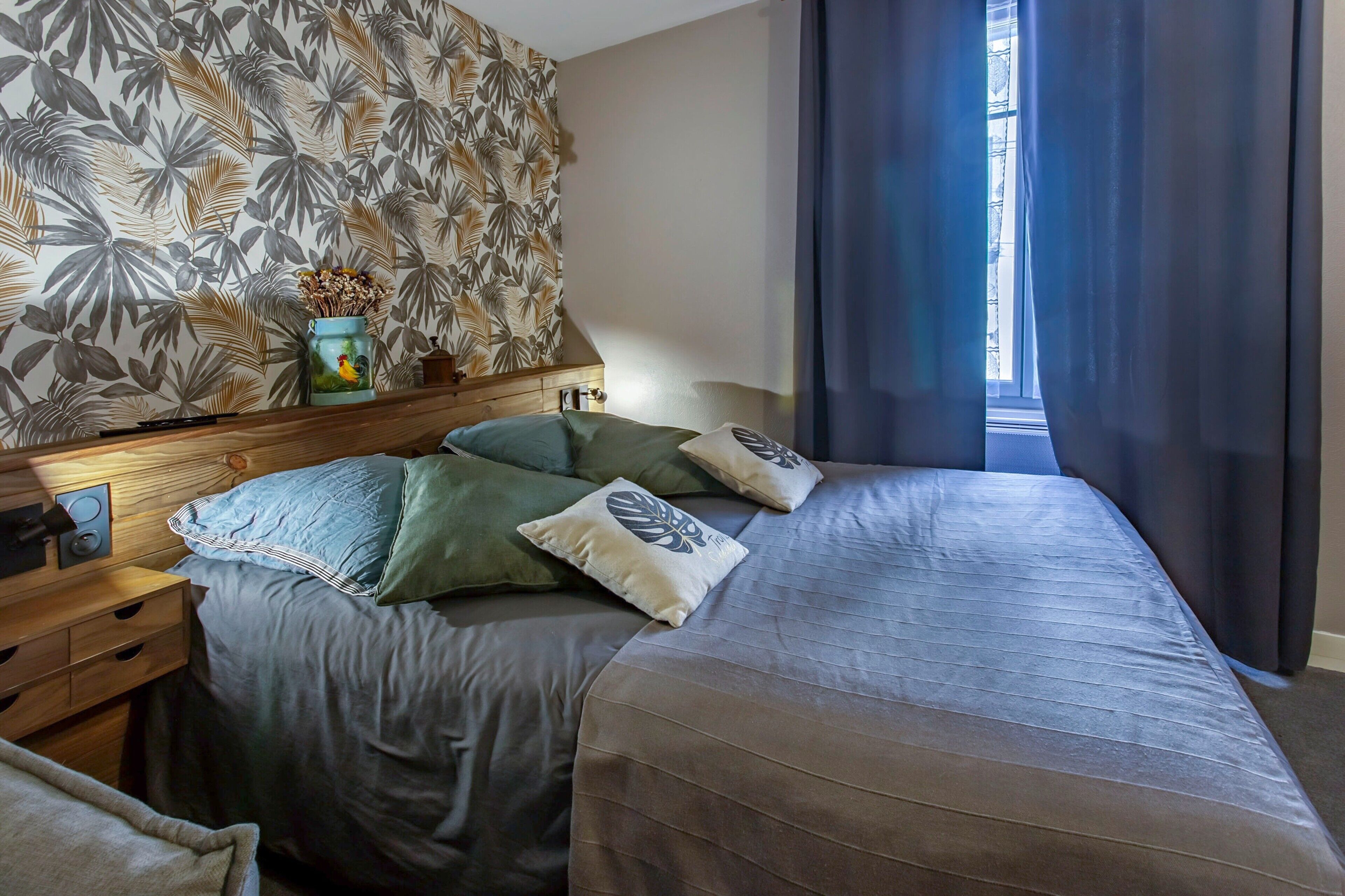 Ein Schlafzimmer mit Bett, Kissen, einem Nachttisch, einer Lampe, einem Fenster mit Vorhängen und einer Wand mit einem Blattmuster.