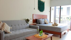Un dormitorio moderno con un sofá gris, una mesita de noche de madera, una cama con colchón gris y un colgador de macramé verde en la pared.