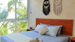 Ein Schlafzimmer mit hölzernem Kopfteil, einem Bett mit blauer Bettwäsche, zwei gerollten Handtüchern und Blick auf Grün durch ein Fenster.