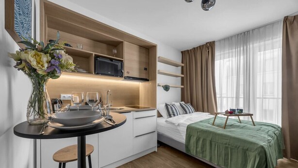 Una camera d'albergo compatta con angolo cottura, zona pranzo, un letto con coperta verde e una finestra con tende.