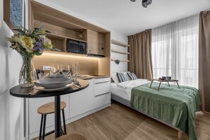 Ein kleines Hotelzimmer mit Küchenzeile, einer Essecke, einem Bett mit grüner Decke und einem Fenster mit Vorhängen.