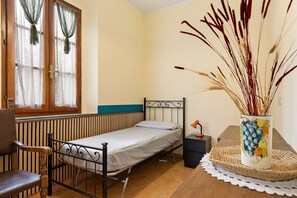 Un lit simple avec une tête de lit en bois, une chaise, une table de chevet avec une lampe et un vase contenant des plantes séchées, le tout posé sur un plateau en osier.