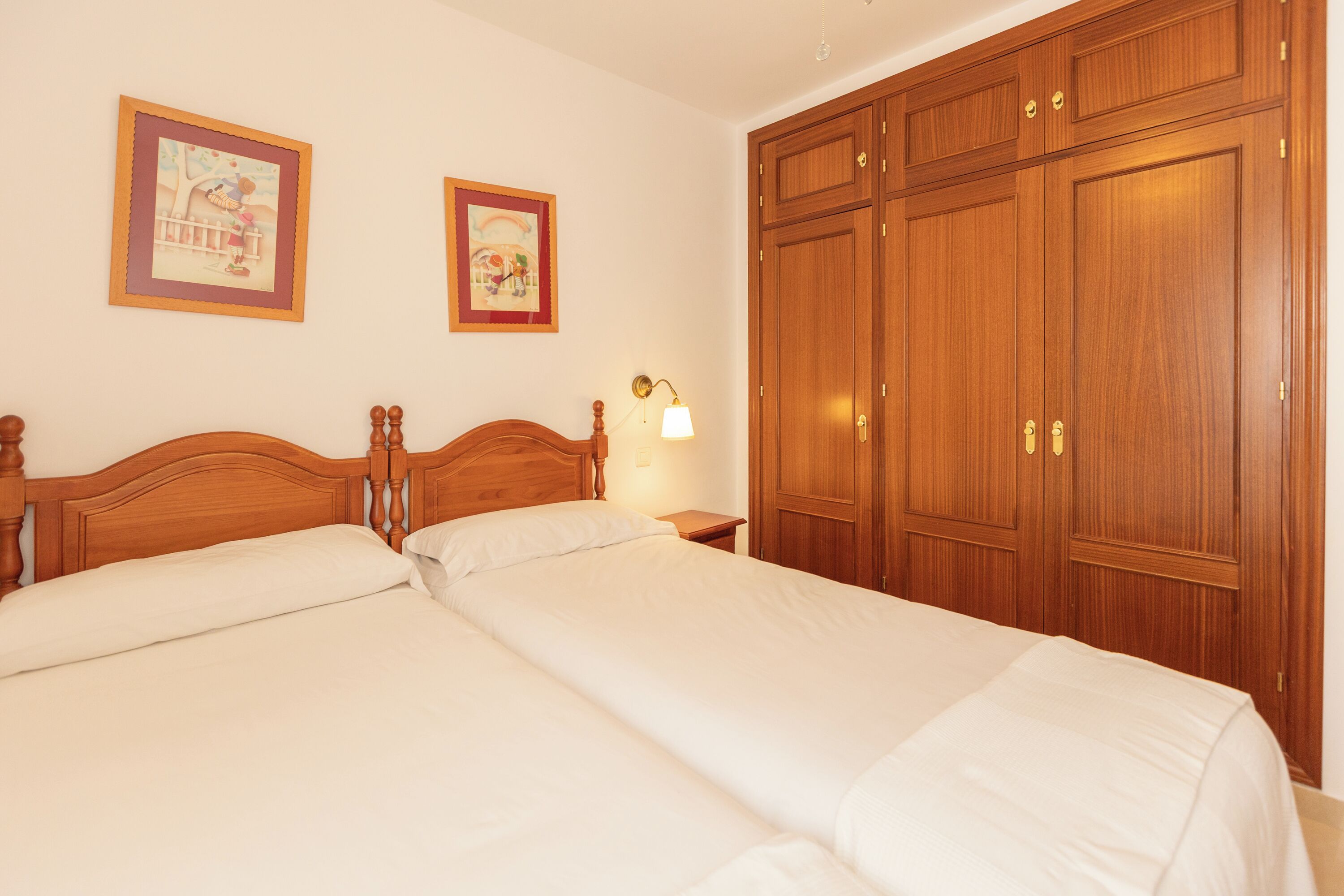 A bedroom with two single beds, wooden wardrobe, and two framed pictures on the wall.