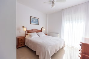 Un dormitorio con cama, mesitas de noche, ventilador de techo, radiador y ventana con cortinas traslúcidas.
