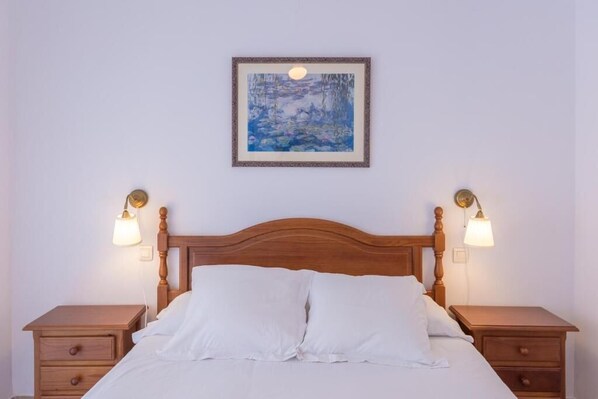 A neatly made bed with white linens, a wooden headboard, and two bedside tables with lamps. - Apartamentos Miguel Ángel (Estepona)