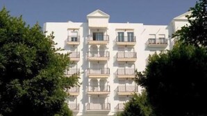Un edificio blanco de varios pisos con balcones, coches aparcados y árboles en la fachada.