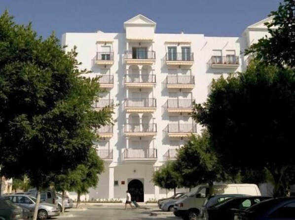A multi-story white building with balconies, parked cars, and trees in front. - Apartamentos Miguel Ángel  (Estepona)
