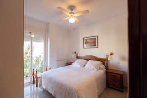Un dormitorio con cama, ventilador de techo, mesita de noche, lámpara, un cuadro en la pared y vistas por la ventana.