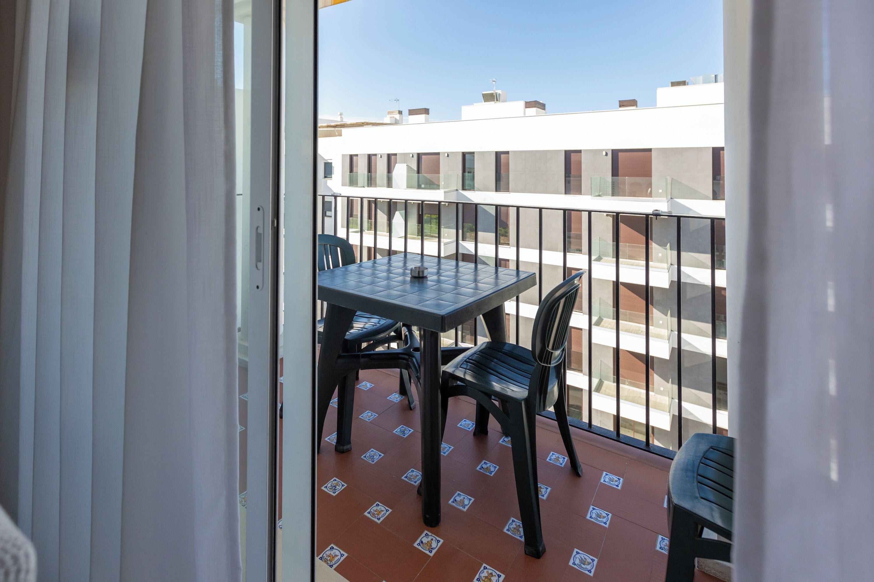 Un balcone con tavolo e sedie, pavimento piastrellato e vista su edifici sullo sfondo.