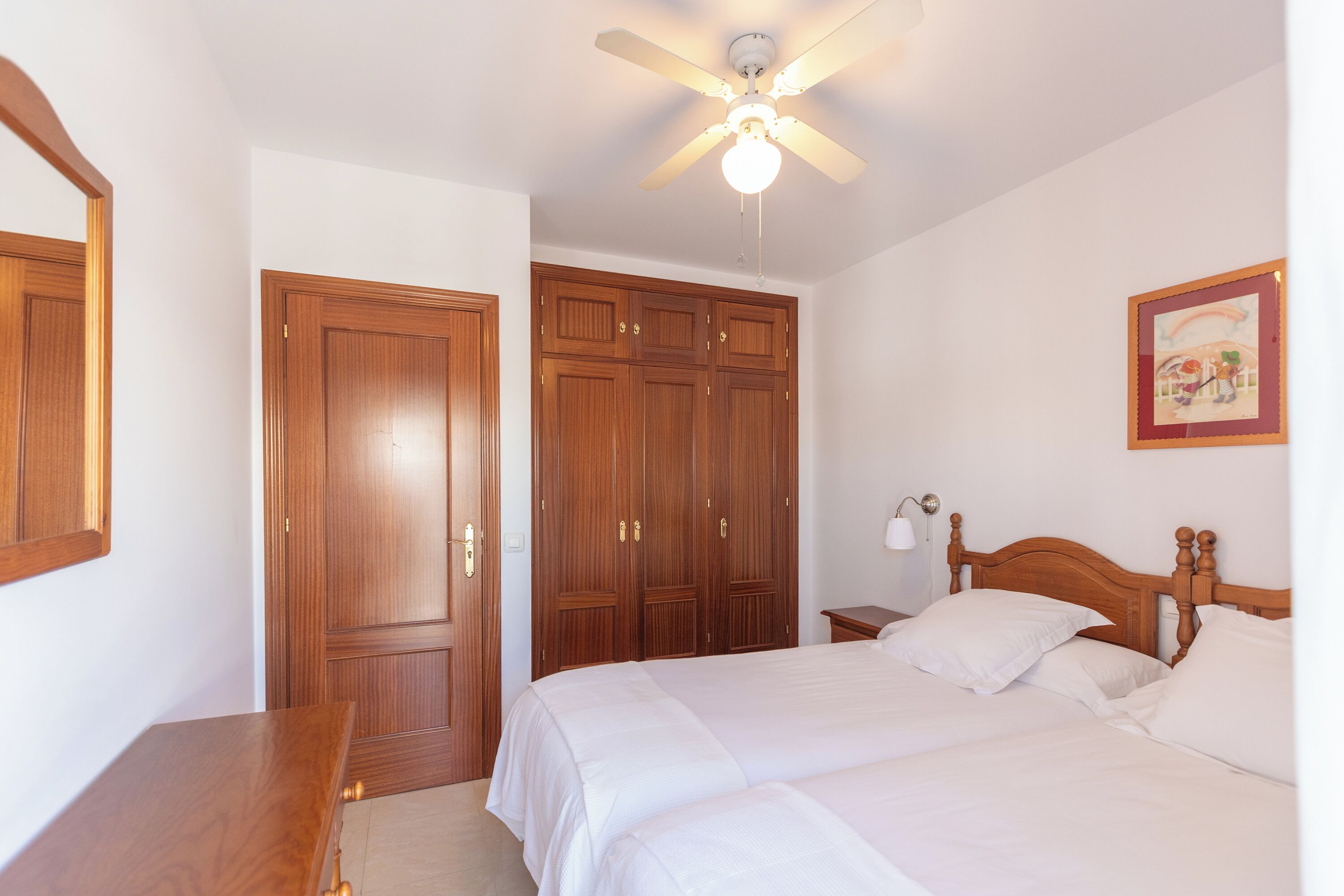 A bedroom with a bed, wooden wardrobe, ceiling fan, and a painting on the wall.