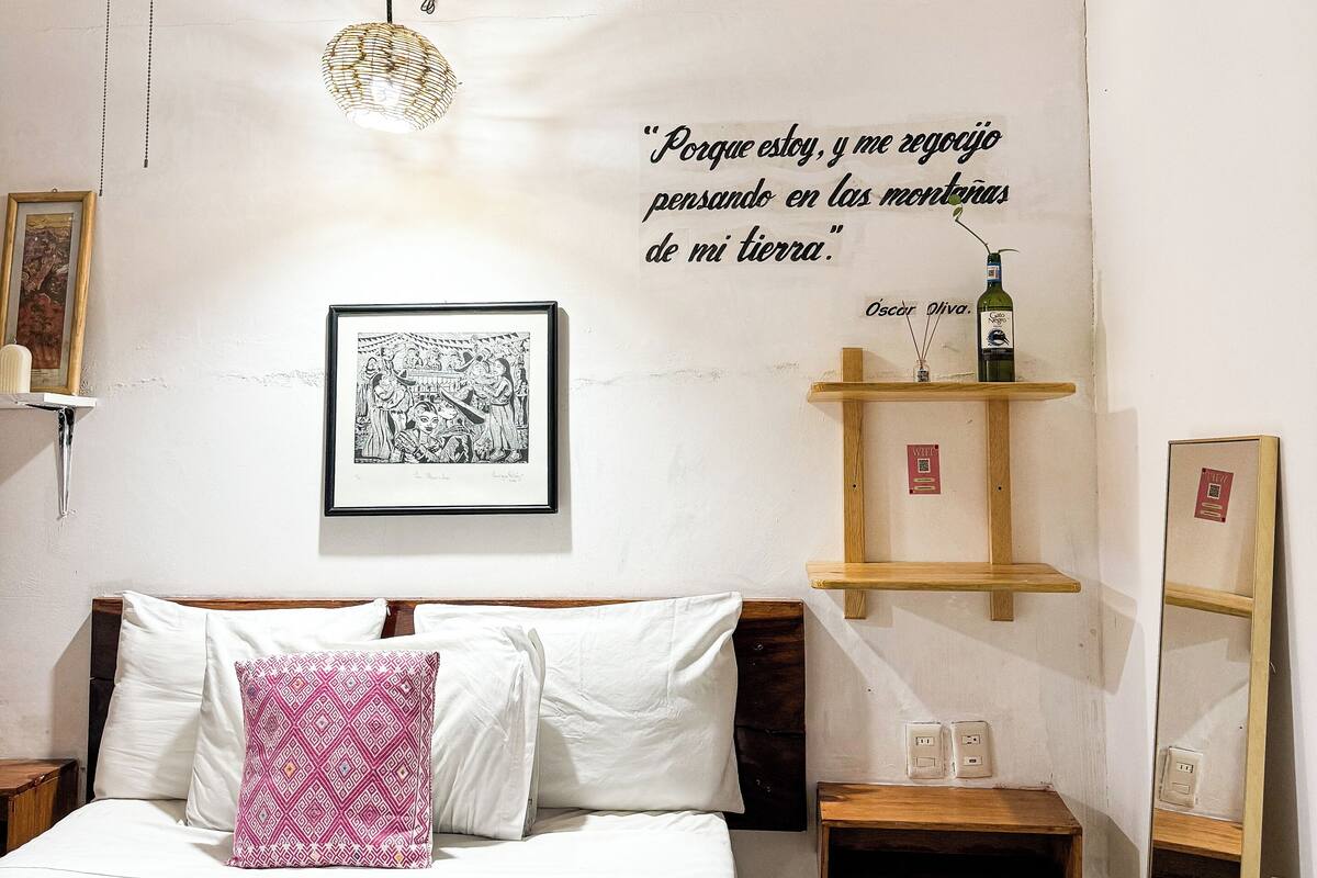 Une chambre à coucher avec un lit, une table de chevet, une étagère avec une bouteille, un tableau encadré et une citation sur le mur.