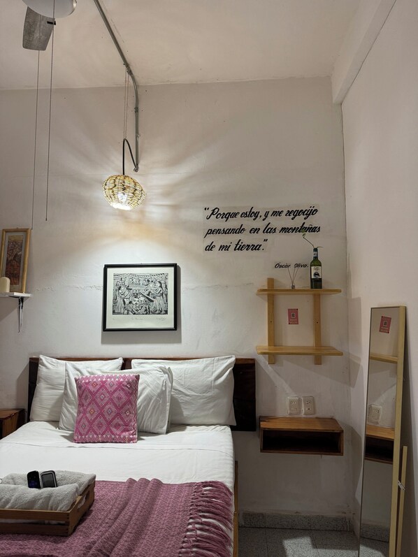 A bedroom with a bed, a bedside table, a shelf with a bottle, and a wall with a quote. - Hostal Casa de Zeferina (Tuxtla Gutiérrez)