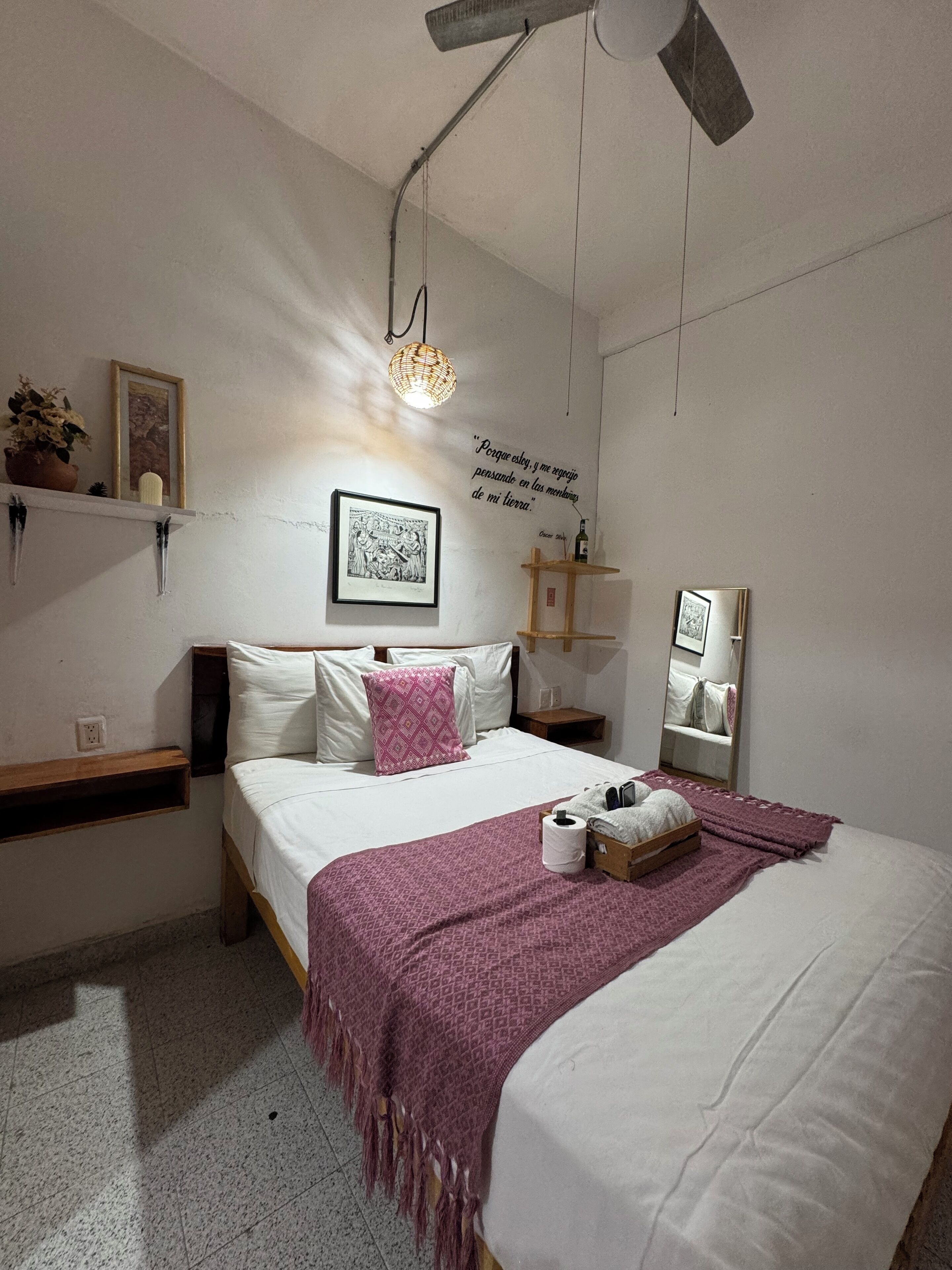 Un dormitorio con cama, mesita de noche, repisa en la pared, lámpara colgante, espejo y una cita decorativa en la pared.