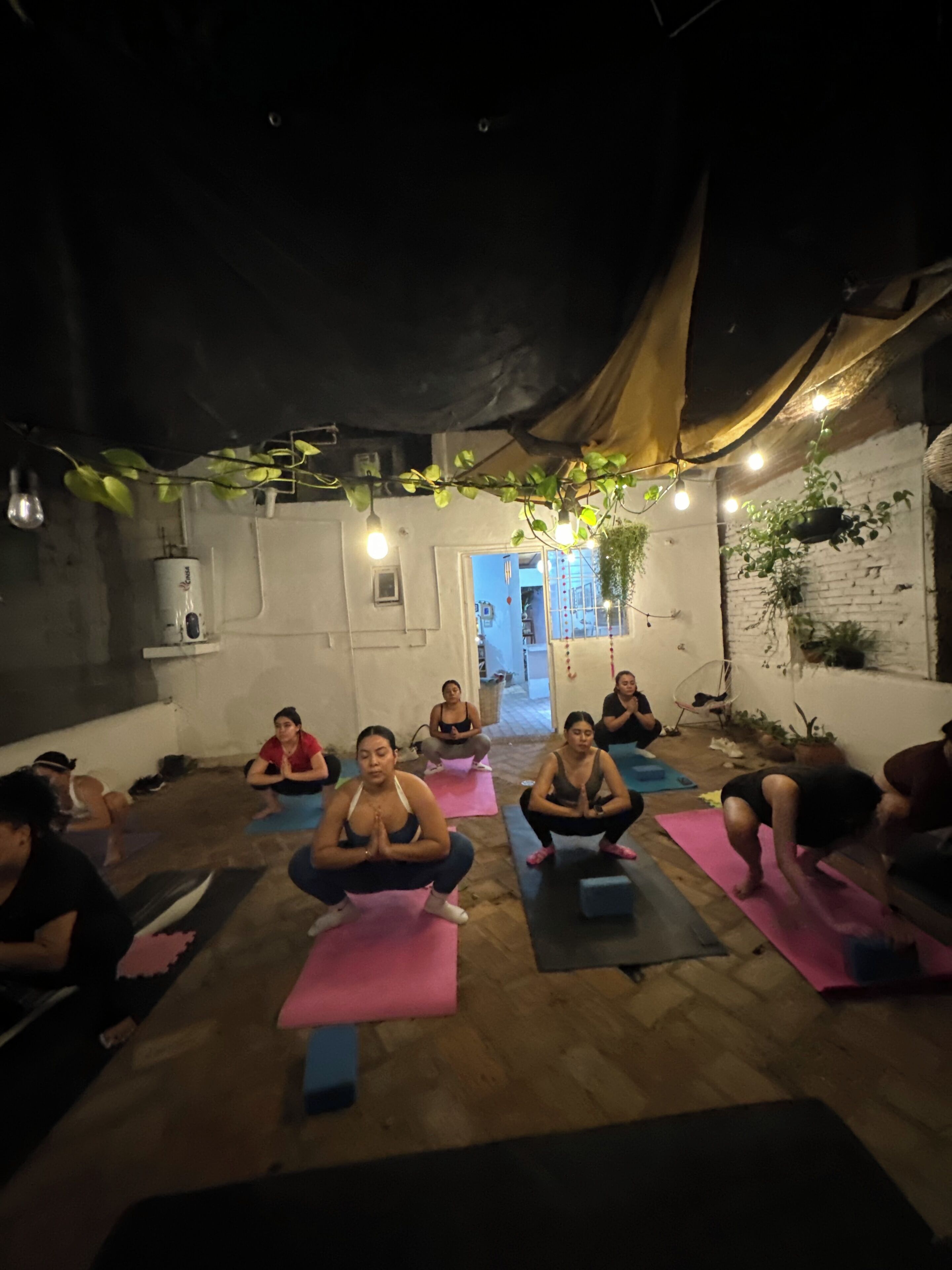 A group of people in a dimly lit room, seated on yoga mats, engaged in a yoga session.