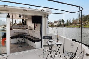 Cubierta de barco con una mesa, sillas, un banco y una botella de champán.