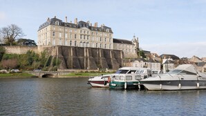 A historic building with a large balcony overlooking a river, boats docked nearby, and a stone bridge in the foreground.