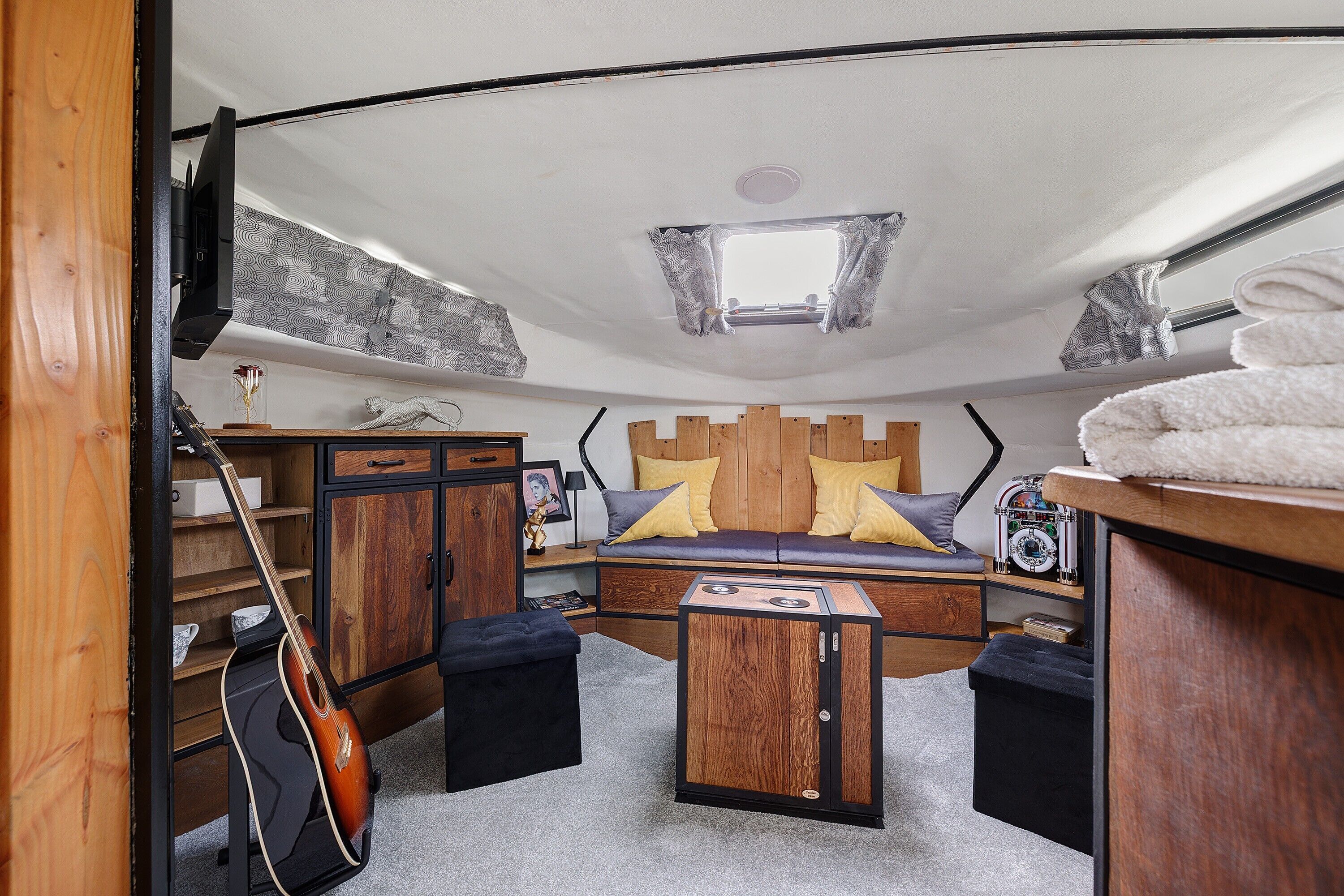 A compact RV interior with a bed, a guitar, a small table, and a cabinet.