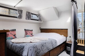 Un dormitorio con una cama, almohadas, una ventana, una cortina y una pequeña mesa con un objeto decorativo.