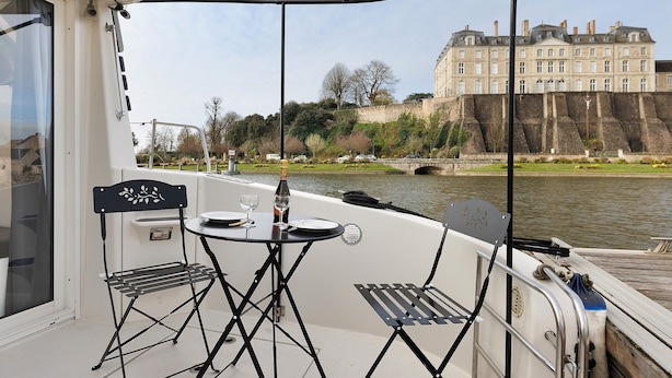 A boat with a table and two chairs, a bottle of wine, and a view of a large building across the water.