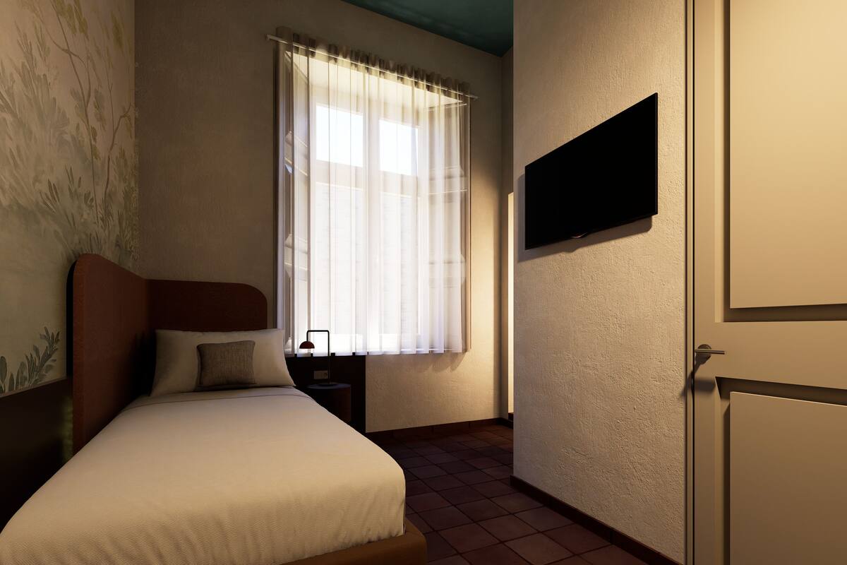 A bedroom with a bed, a nightstand, a TV mounted on the wall, and a window with sheer curtains.