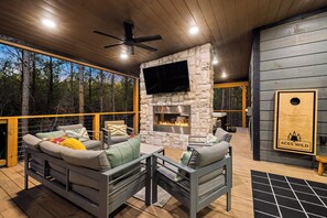 A cozy outdoor seating area with a stone fireplace, a flat-screen TV mounted above it, and a wooden deck with a railing. - Luxe & Kid Approved! Playset, EV, Meta VR Game (Broken Bow)