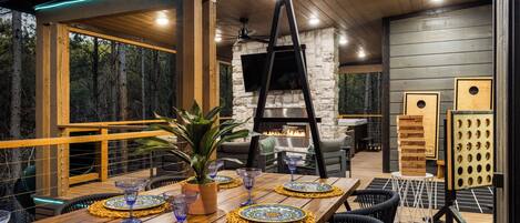 Una terraza de madera con una mesa de comedor dispuesta para seis personas, rodeada de sillas. Se ve una chimenea de piedra al fondo y hay macetas de madera con plantas en la terraza.