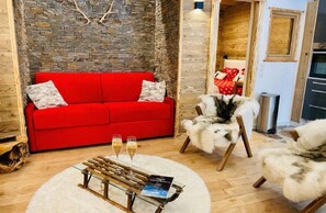 Un salon chaleureux avec un canapé rouge, un fauteuil blanc à poils longs et une table basse en verre sur laquelle reposent un livre et deux verres de vin.