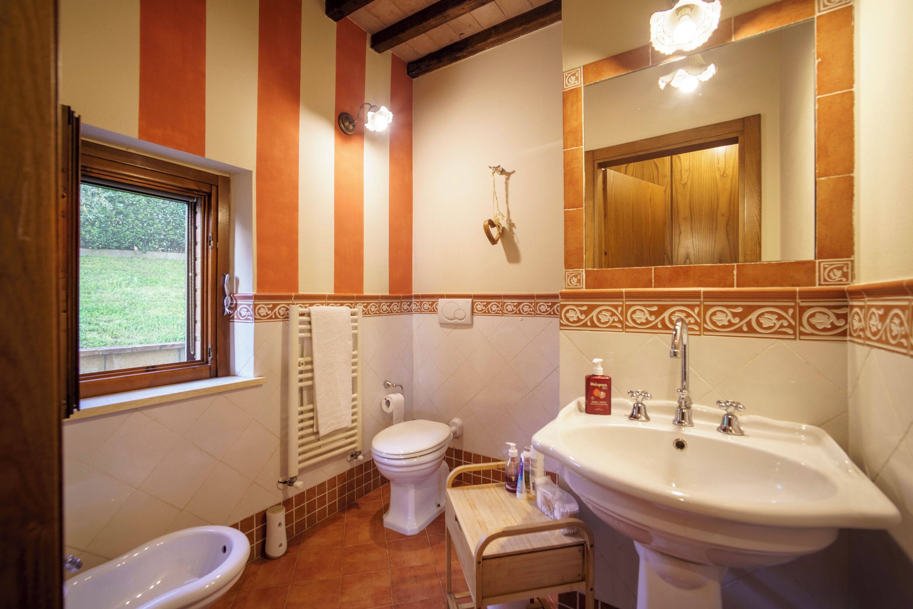 A bathroom with a toilet, bidet, sink, and a window with a view of greenery outside.