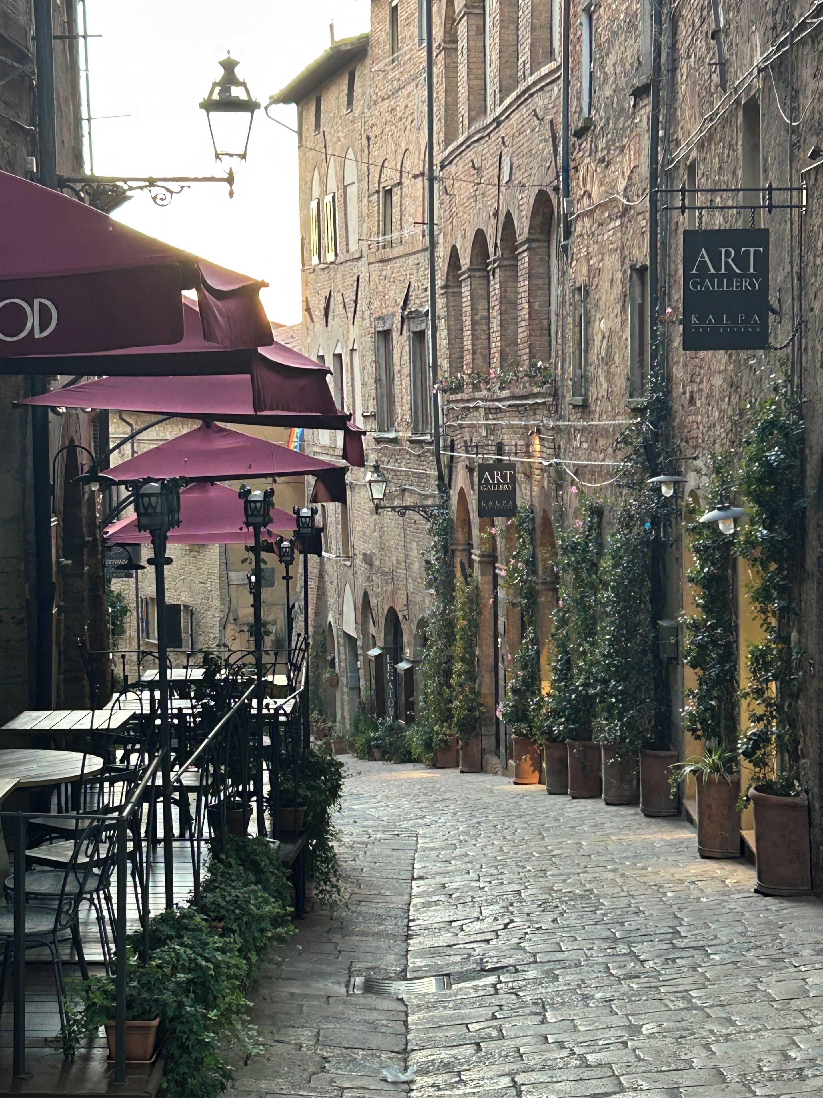 A cobblestone street with outdoor seating, stone buildings, and hanging signs for an art gallery.