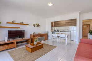A modern living room with a flat-screen TV, wooden entertainment unit, and a red sofa. A kitchen with white cabinets and a dining area with white chairs is visible in the background. - #370 1BR Riamar | Sea View & Central (Armação de Pêra)