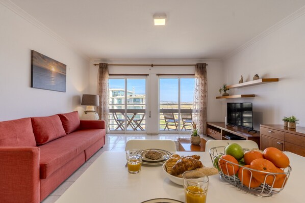 A modern living room with a red sofa, dining table with breakfast items, a TV, and a balcony with a city view. - #370 1-Bedroom Riamar | Ocean View & Central Location (Armação de Pêra)