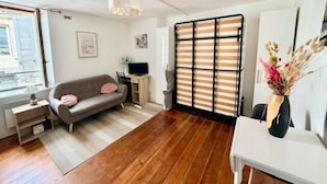 Ein Zimmer mit einer Couch, einem kleinen Tisch, einem Fernseher, einem Schreibtisch und einem Fenster mit Jalousien.