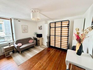 A room with a sofa, a small table, a TV, a desk, and a window with blinds. - Bright studio in the heart of the Old Market (La Rochelle)
