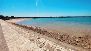 Ein gepflasterter Weg entlang ruhigem Gewässer, blauer Himmel darüber.