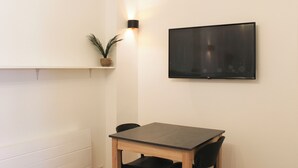 Ein Zimmer mit Holztisch, zwei schwarzen Stühlen, einem fest montierten Flachbildfernseher, einer weißen Wand und einem Regal mit einer Pflanze.