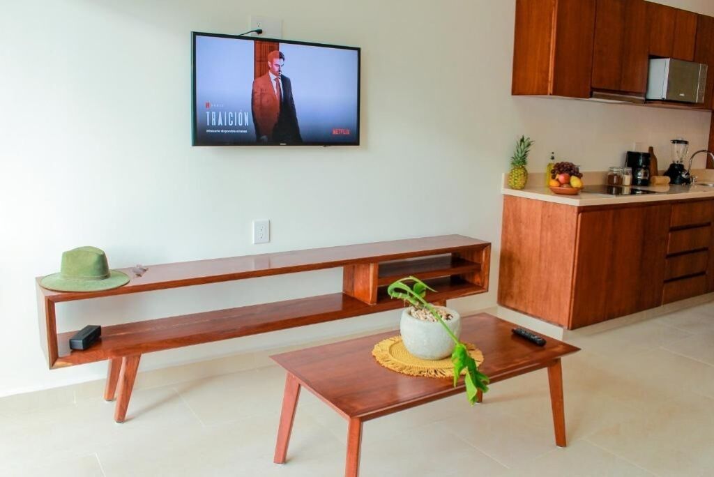 A modern living room with a TV mounted on the wall, a wooden bench, a coffee table with a plant, and a kitchen area with wooden cabinets and a fruit bowl.