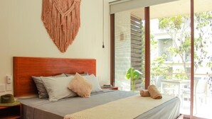 A bedroom with a wooden headboard, a bed with grey bedding, a macrame wall hanging, and a view of a balcony with outdoor furniture.