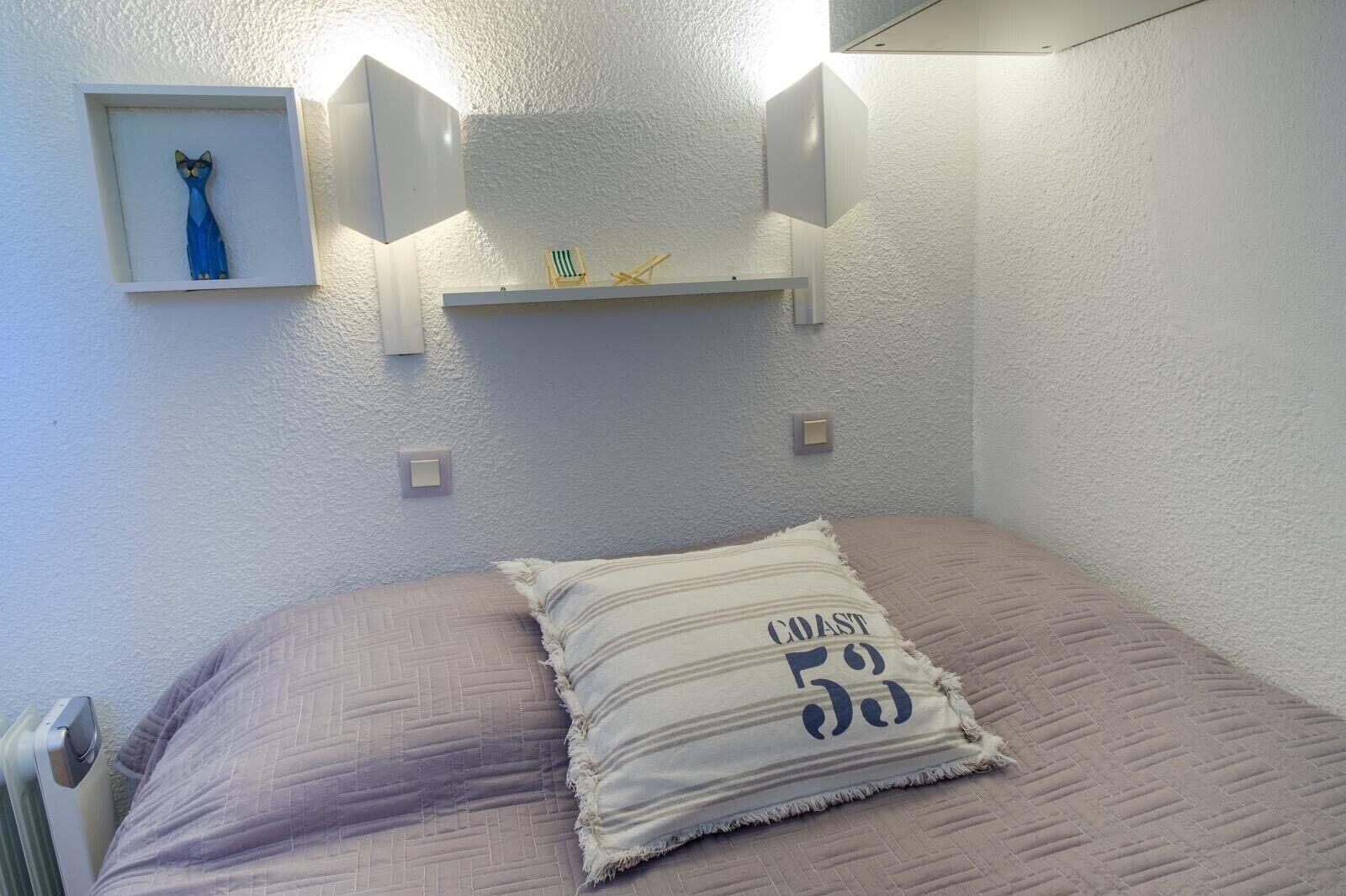 A bedroom with a bed, a pillow with text, a shelf with a small item, and wall-mounted lights.
